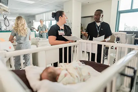 Two healthcare providers talking with a baby in a cot in the foreground