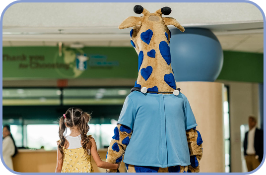 George the Giraffe walks hand in hand with a young girl in the hospital